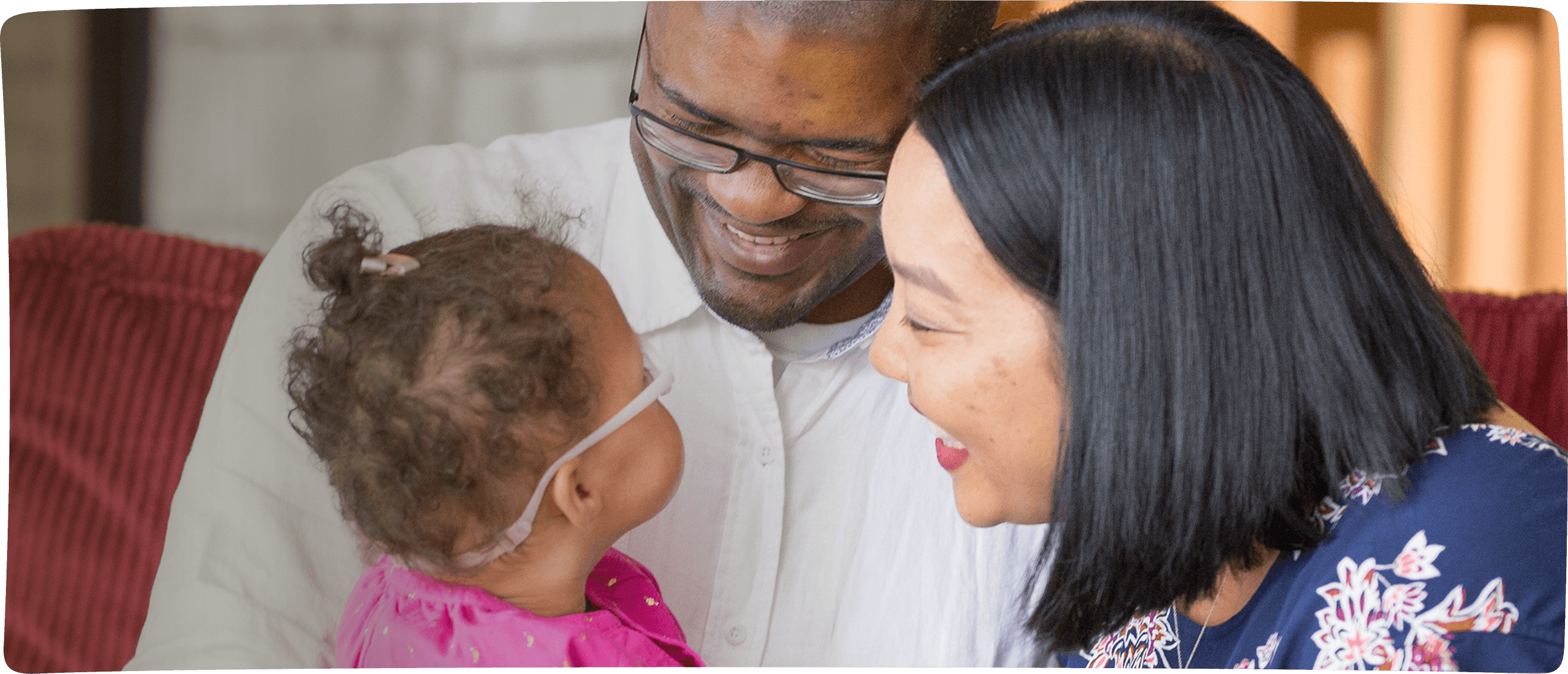 An RMHC family, the Burrells, left to right, infant Piper smiled at by dad Kevin and mom Lindsay, sitting on a red couch, a gentle fire in the fireplace in soft focus behind them