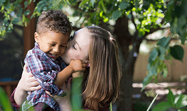 An Ronald McDonald House family, mom Rachel Jolivard holding son Jacob by the waist and kissing his cheek, both enjoying the sun