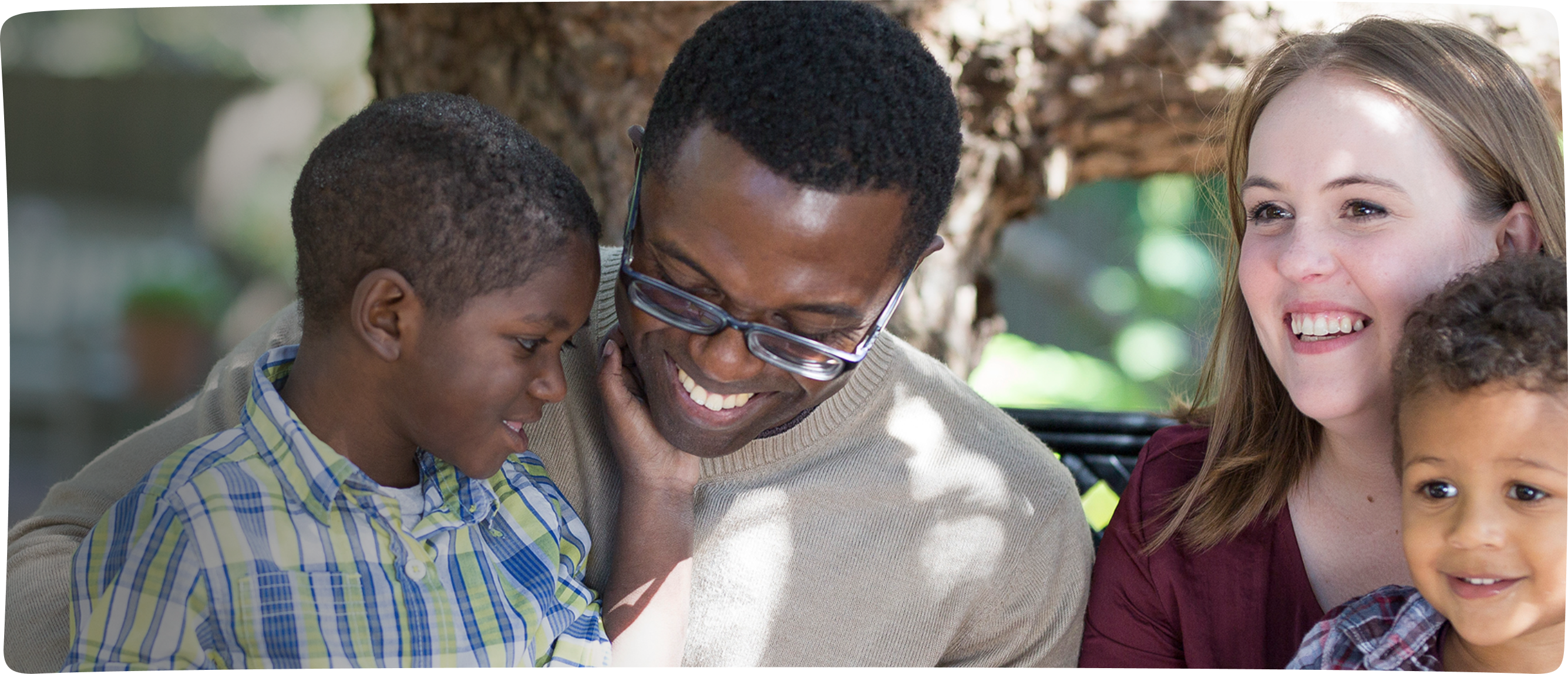 The Jolivards, an RMHC family, Daniel cups the chin of dad Junior who holds him on his lap, mom Rachel holds younger brother Jacob on her lap, all smiling on a sunny day in the shade of a tree
